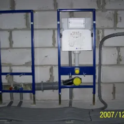 Stelaż podtynkowy Geberit Duofix Basic zamontowany przy ścianie z betonu komórkowego, widoczne rury doprowadzające wodę i odpływ kanalizacyjny, oraz przewody grzewcze w podłodze.