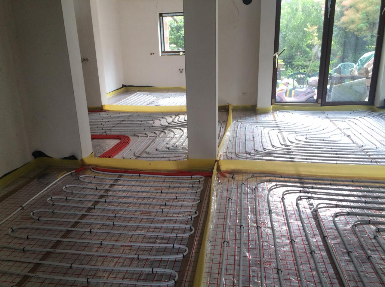 Rozłożona instalacja ogrzewania podłogowego w trakcie montażu, widoczne białe rury na folii z czerwoną siatką, zabezpieczone żółtą taśmą na obrzeżach pomieszczenia.