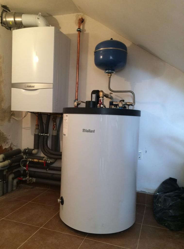 Kocioł gazowy Vaillant wiszący obok pionowego zbiornika na wodę, z widocznymi rurami miedzianymi i szarymi rurami kanalizacyjnymi, w pomieszczeniu z brązowymi płytkami.