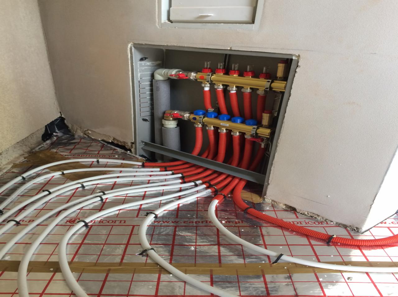 Rozdzielacz ogrzewania podłogowego z rurami doprowadzającymi, widoczne białe i czerwone rury na folii izolacyjnej z nadrukiem, zamontowany w ścianie, surowy stan wykończenia.