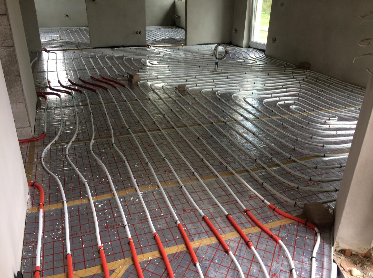 Rozłożona instalacja ogrzewania podłogowego w surowym wnętrzu, rury grzewcze ułożone spiralnie na folii z siatką, widoczne fragmenty ścian i okna.