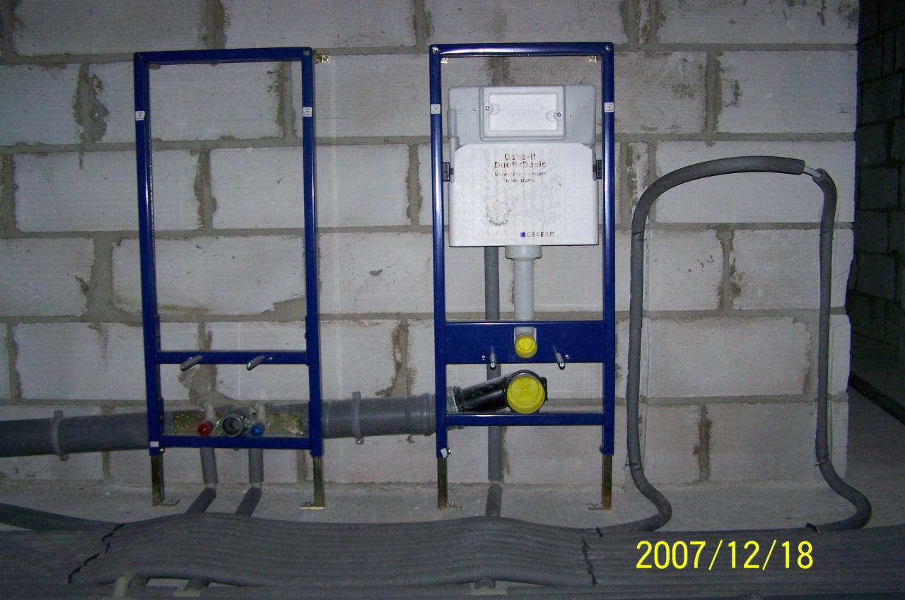 Stelaż podtynkowy Geberit Duofix Basic zamontowany przy ścianie z betonu komórkowego, widoczne rury doprowadzające wodę i odpływ kanalizacyjny, oraz przewody grzewcze w podłodze.