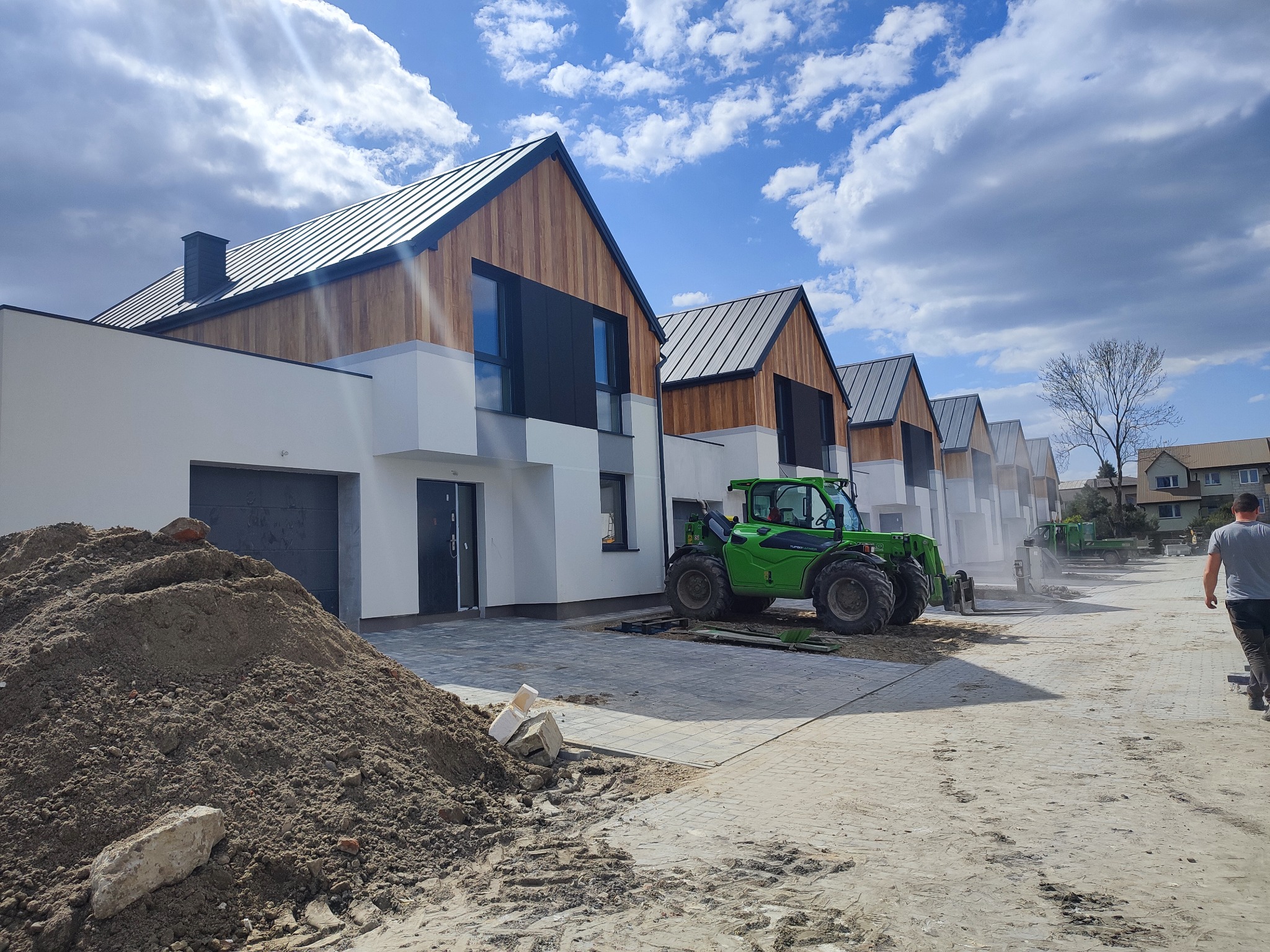Szereg nowoczesnych domów jednorodzinnych z drewnianą elewacją i ciemnymi dachami, w trakcie budowy; widoczny zielony wózek widłowy i sterta ziemi.