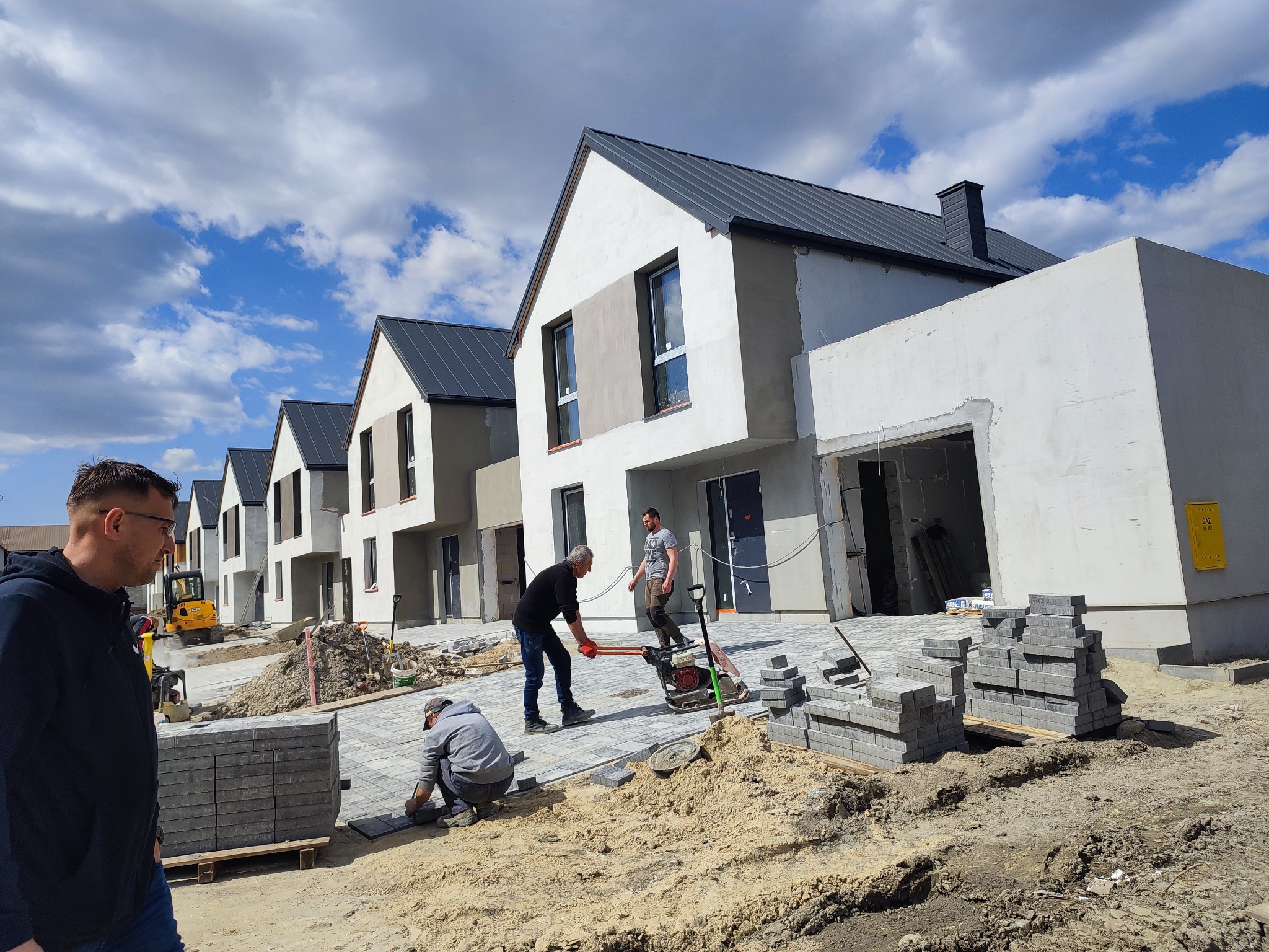 Budowa osiedla domów jednorodzinnych z szarymi dachami, robotnicy układają kostkę brukową na podjeździe, widoczna ubijarka i stosy kostki.