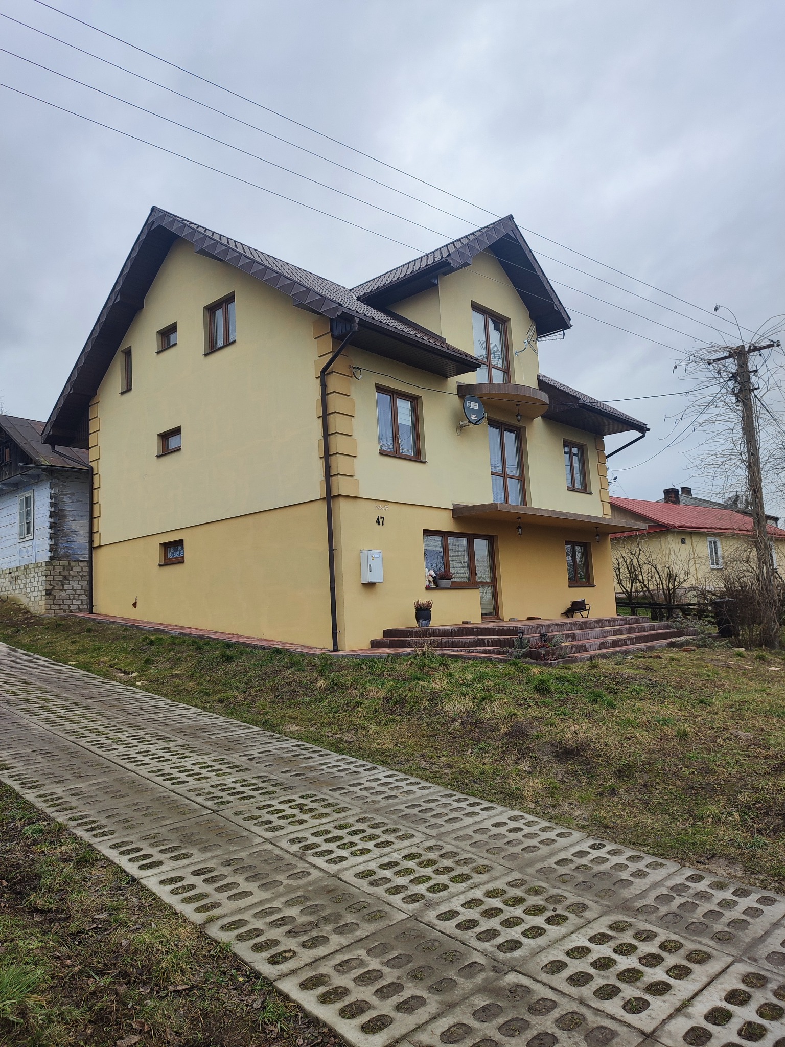 Dwupiętrowy dom z żółtą elewacją, brązowym dachem i balkonem, widziany z dołu ścieżki z betonowych płyt ażurowych.