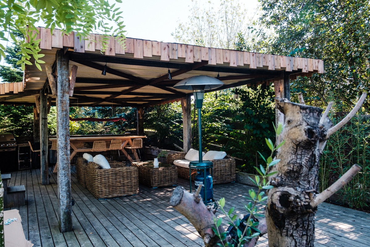 Zadaszony taras z drewnianą podłogą i meblami rattanowymi, oświetlony lampą grzewczą, z widokiem na bujną zieleń ogrodu.