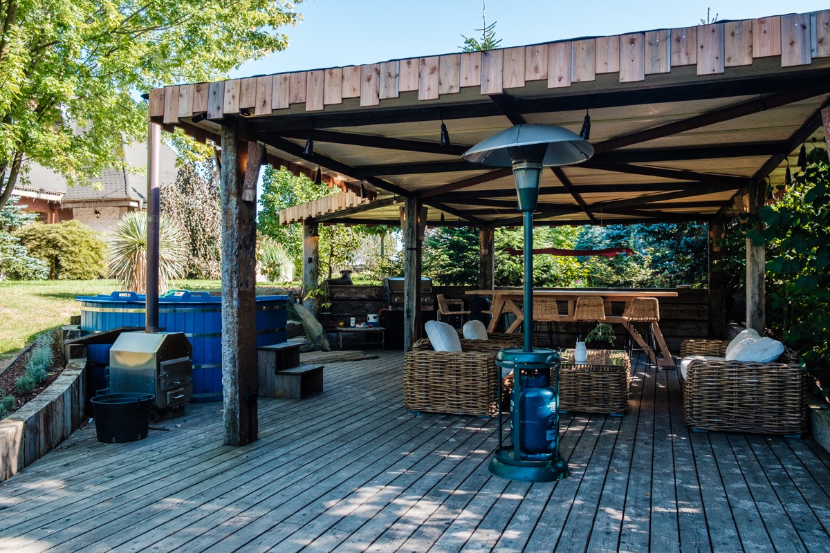 Zadaszony taras z drewnianą podłogą, rattanowymi meblami, zewnętrznym jacuzzi w kolorze niebieskim i lampą grzewczą na gaz w słoneczny dzień.