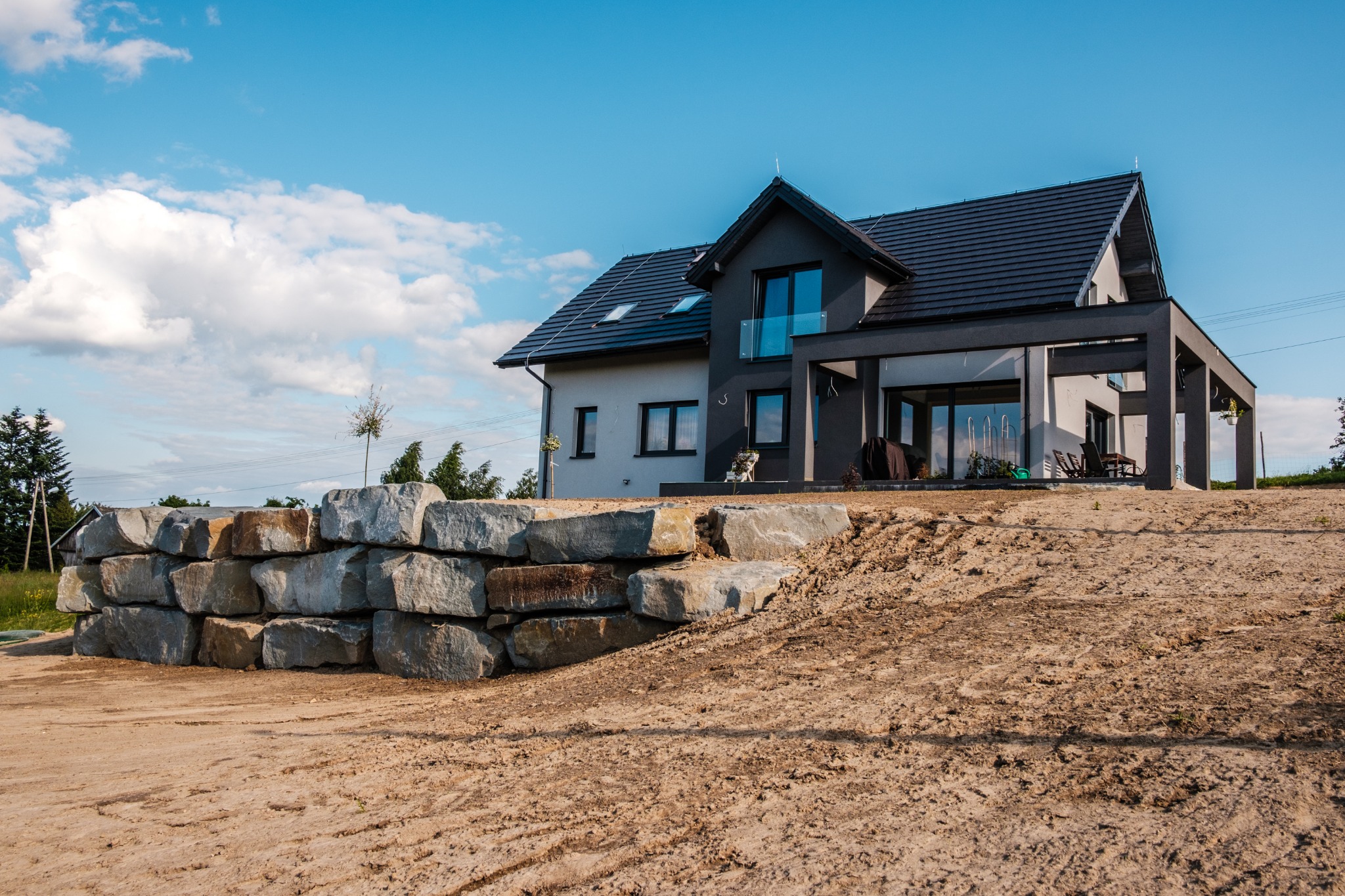 Nowoczesny dom z szarą elewacją i dużymi oknami, otoczony ziemią w trakcie prac ziemnych, z kamiennym murem oporowym z szarego kamienia.