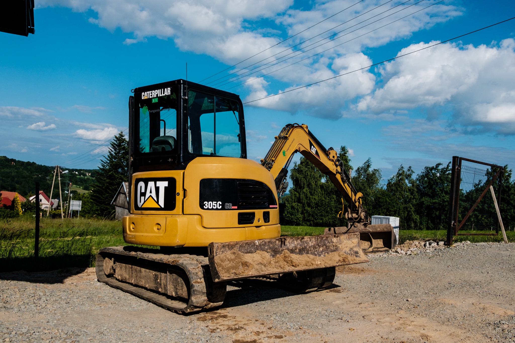 Żółta minikoparka Caterpillar 305C CR na żwirowym podłożu, z widocznym lemieszem zabrudzonym ziemią, na tle błękitnego nieba z chmurami i fragmentami zieleni.