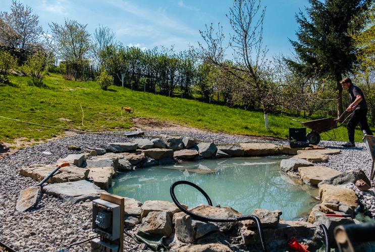 Budowa oczka wodnego: pracownik z taczką, wykop wyłożony kamieniami, woda w zbiorniku, narzędzia i skrzynka elektryczna w pobliżu.