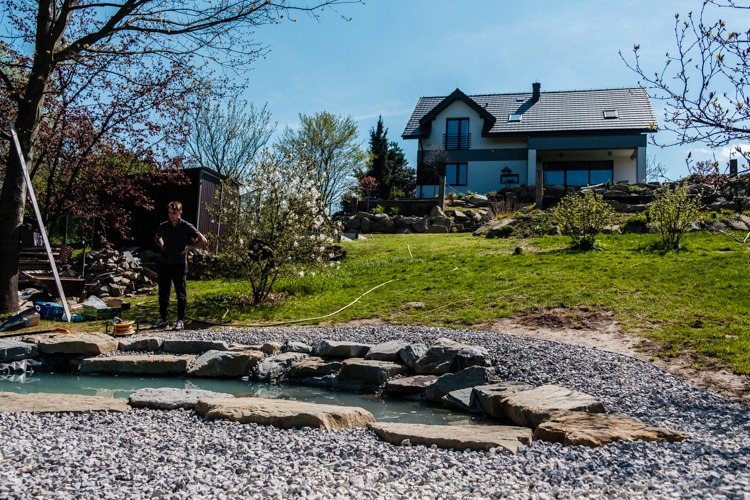 Budowa strumienia z kamieni w ogrodzie, pracownik nadzoruje postęp prac, w tle dom jednorodzinny i zieleń.