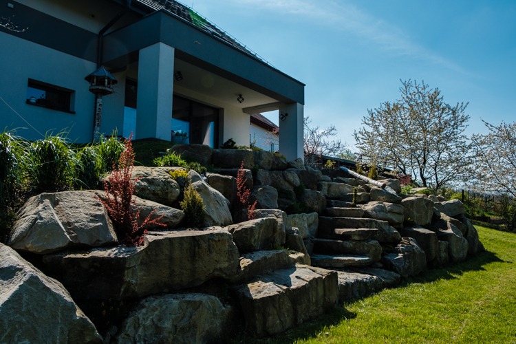 Ogród skalny z kamiennymi schodami prowadzącymi do domu, z nasadzeniami roślin ozdobnych, na tle nowoczesnej architektury i błękitnego nieba.