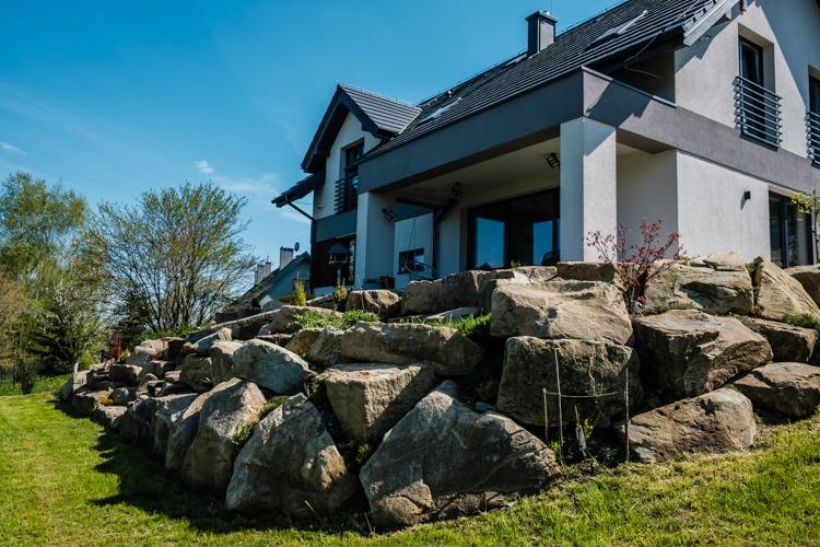 Skarpa z dużych, nieregularnych kamieni na tle nowoczesnego domu z szarym dachem i białymi ścianami, zielony trawnik na pierwszym planie, błękitne niebo w tle.