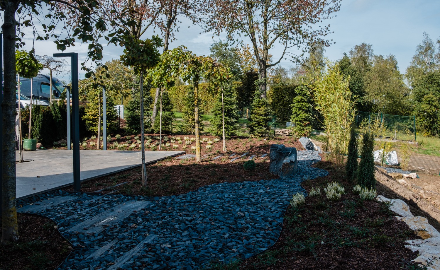 Nowoczesny ogród z ścieżką z ciemnych kamieni, drzewami formowanymi, podwyższonym tarasem i ozdobnymi krzewami.