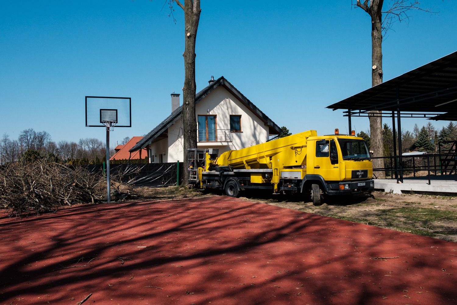 Żółty podnośnik koszowy MAN na tle domu jednorodzinnego i boiska do koszykówki po wycince drzew, z gałęziami na ziemi.