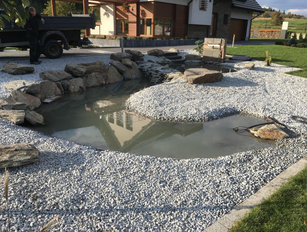 Nowo wybudowane oczko wodne z kamieniami i białym żwirem, odbicie domu w tafli wody, widoczny wąż i sprzęt budowlany.