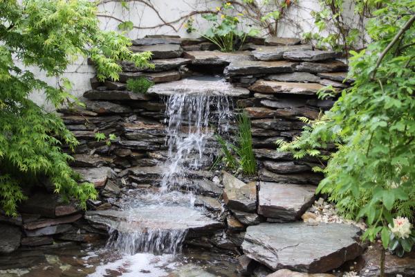 Niewielki wodospad kaskadowy z ciemnego kamienia łupkowego, otoczony zielenią, z roślinami pnącymi się po jasnej ścianie w tle.