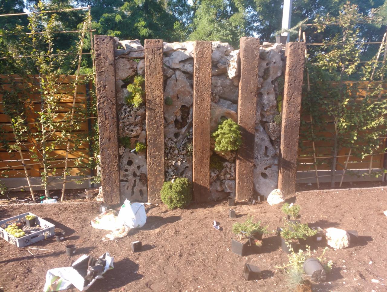 Wertykalny ogród w trakcie tworzenia: kamienna ściana z drewnianymi belkami, nasadzenia roślin w szczelinach, rozłożone narzędzia i sadzonki na ziemi.