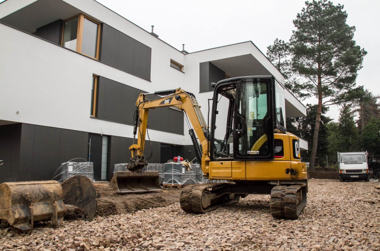 Żółta minikoparka CAT na żwirowym podłożu przed nowoczesnym domem z czarno-białą elewacją, przygotowująca teren pod przyszły ogród.