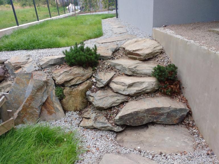 Kamienne schody w ogrodzie z niskimi iglakami i żwirem, prowadzące wzdłuż ściany budynku.