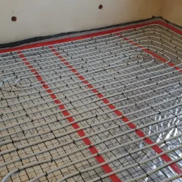 Rozłożona instalacja ogrzewania podłogowego w Gdyni, widoczne białe rurki na czarnej siatce i czerwone taśmy mocujące na folii aluminiowej, przygotowane do zalania wylewką.