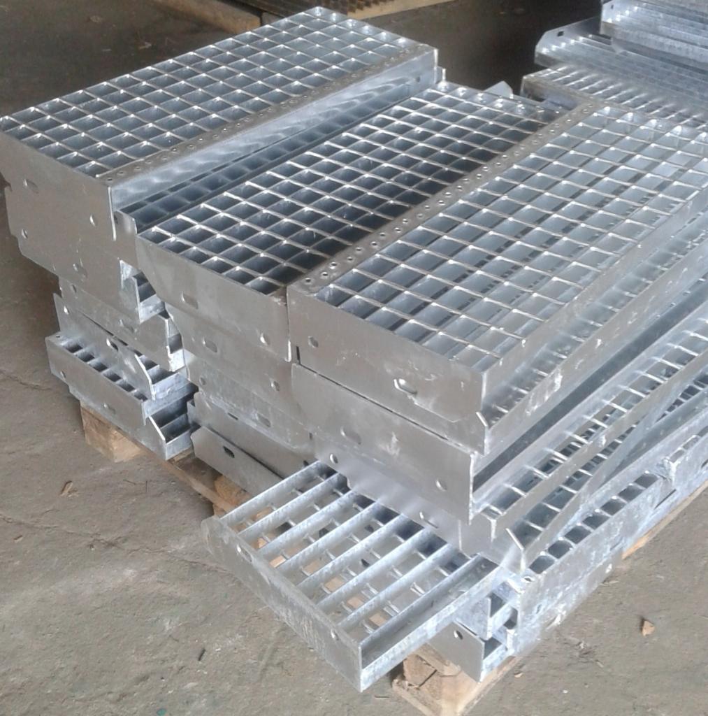Stos ocynkowanych, kratowych podestów stalowych na drewnianej palecie, widoczne otwory montażowe i spawane łączenia.