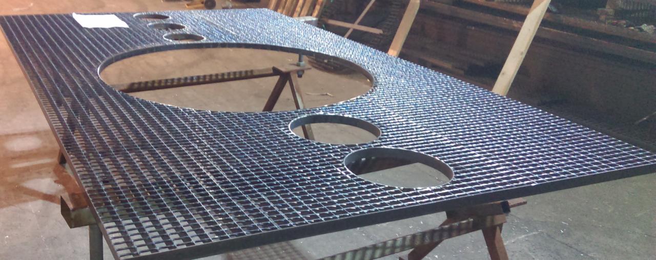 Metalowa krata pomostowa z okrągłymi otworami, spawana konstrukcja na metalowych kozłach w hali produkcyjnej.