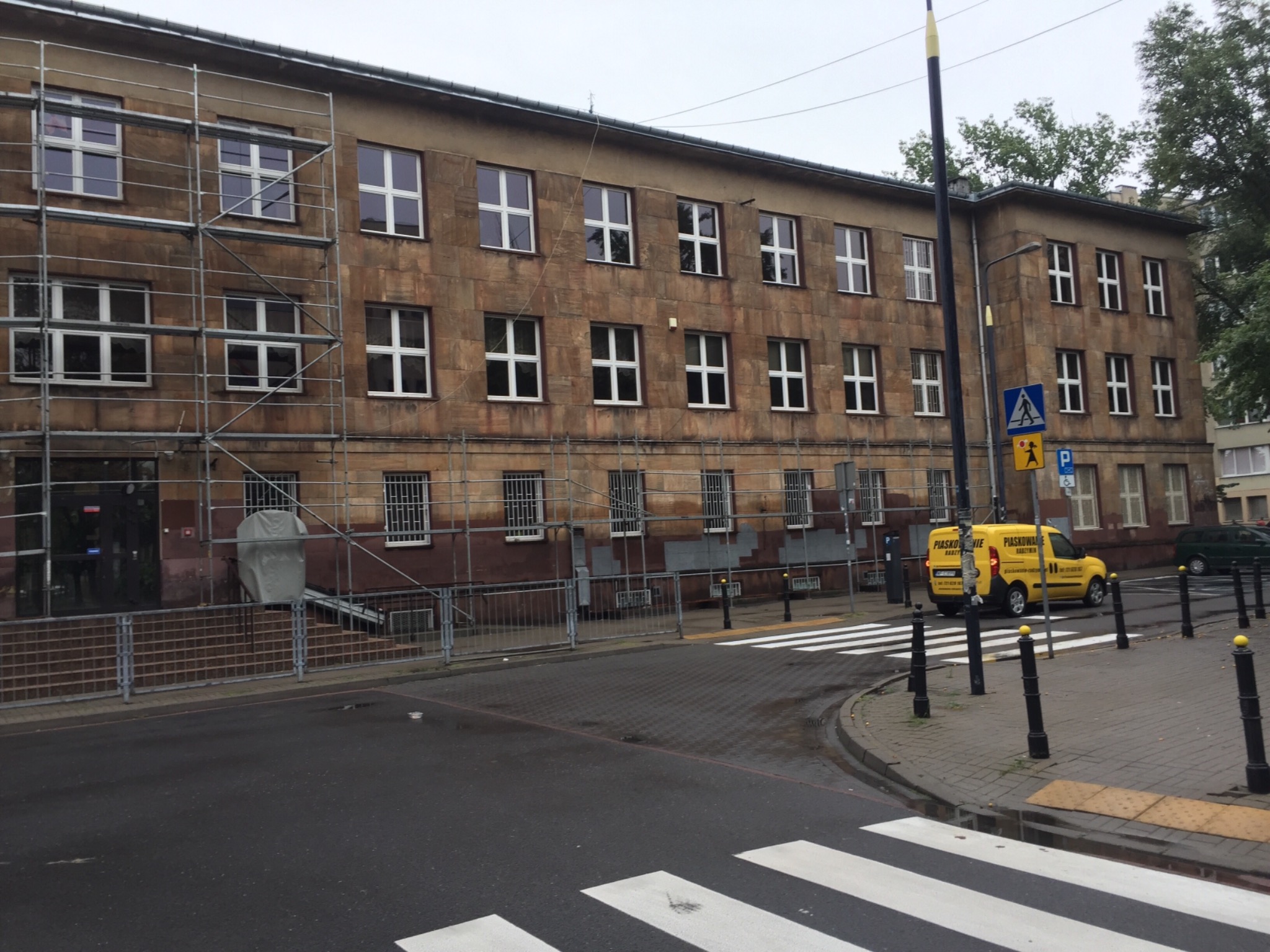 Budynek z piaskowca w trakcie renowacji, widoczne rusztowania, żółty samochód dostawczy z napisem na boku, przejście dla pieszych i słupki.