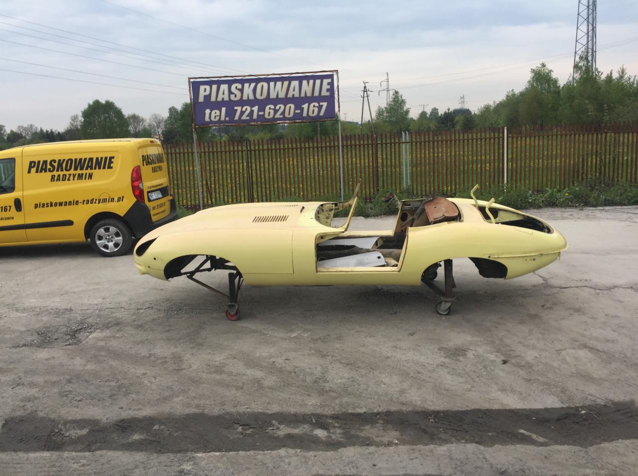Żółta karoseria klasycznego samochodu Jaguar E-Type, przygotowana do renowacji, na tle reklamy firmy i samochodu dostawczego.