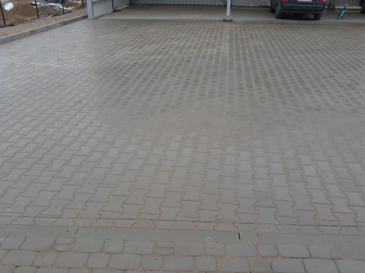 Szara kostka brukowa ułożona we wzór jodełki na podjeździe, widoczne szczeliny wypełnione piaskiem, w tle wiata garażowa i zaparkowany samochód.
