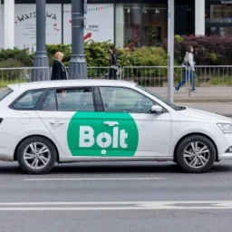 Biała Skoda Fabia kombi z zielonym logo Bolt na drzwiach, jadąca po pasach na tle miejskiego krajobrazu z pieszymi.