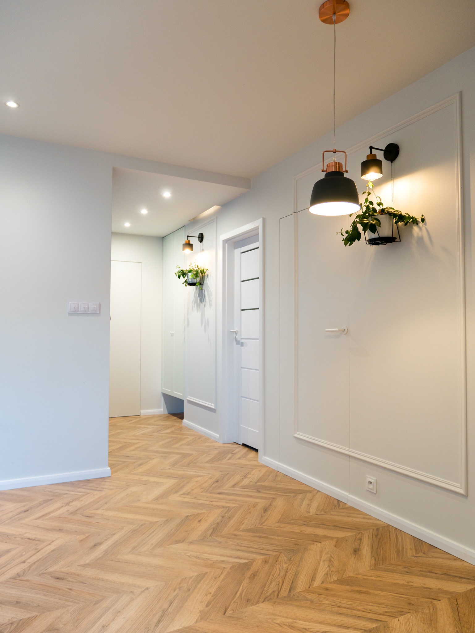 Jasne wnętrze: parkiet w jodełkę, białe ściany z dekoracyjną listwą, drzwi z przeszkleniami i oświetlenie punktowe oraz wiszące lampy z roślinami.