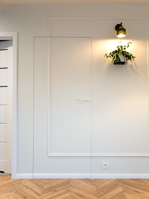 Biała ściana z ukrytymi drzwiami i dekoracyjną sztukaterią. Na ścianie kinkiet oświetlający roślinę doniczkową. Parkiet ułożony w jodełkę.