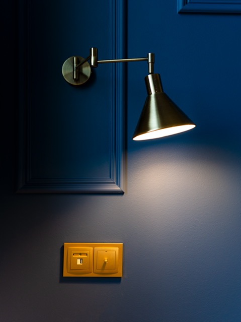 Granatowa ściana z dekoracyjną ramą, oświetlona złotą lampą ścienną i gniazdkiem z włącznikiem w stylu retro. Subtelne światło tworzy ciepły klimat.