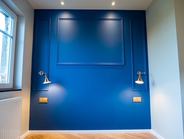 Niebieska ściana z dekoracyjnymi listwami i złotymi kinkietami. Parkiet ułożony w jodełkę. Nowoczesne wykończenie wnętrza z dbałością o detale.