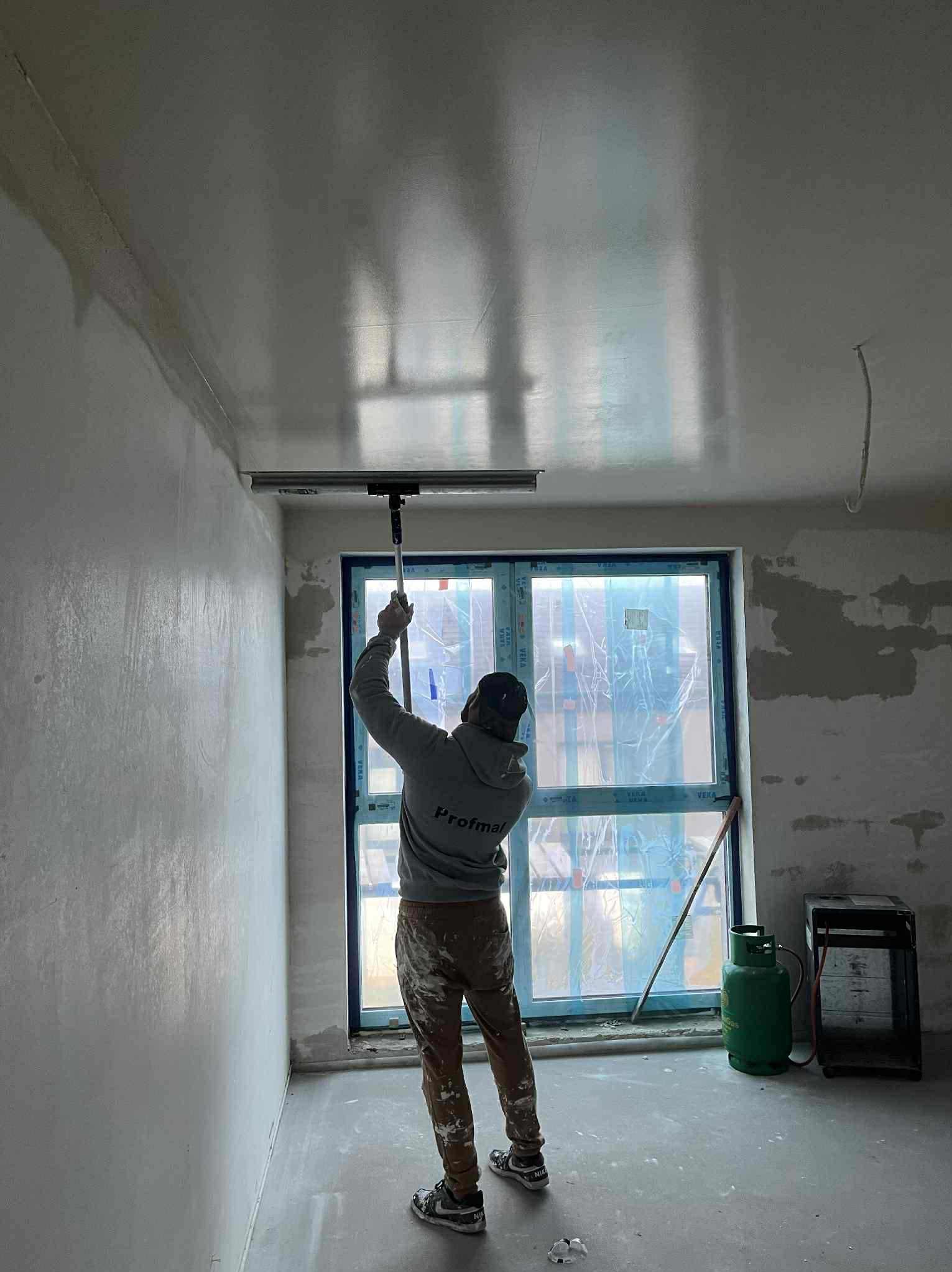 Pracownik w trakcie nakładania gładzi szpachlowej na sufit za pomocą specjalnej pacy. Widoczne okno z folią ochronną i butla gazowa w pomieszczeniu.