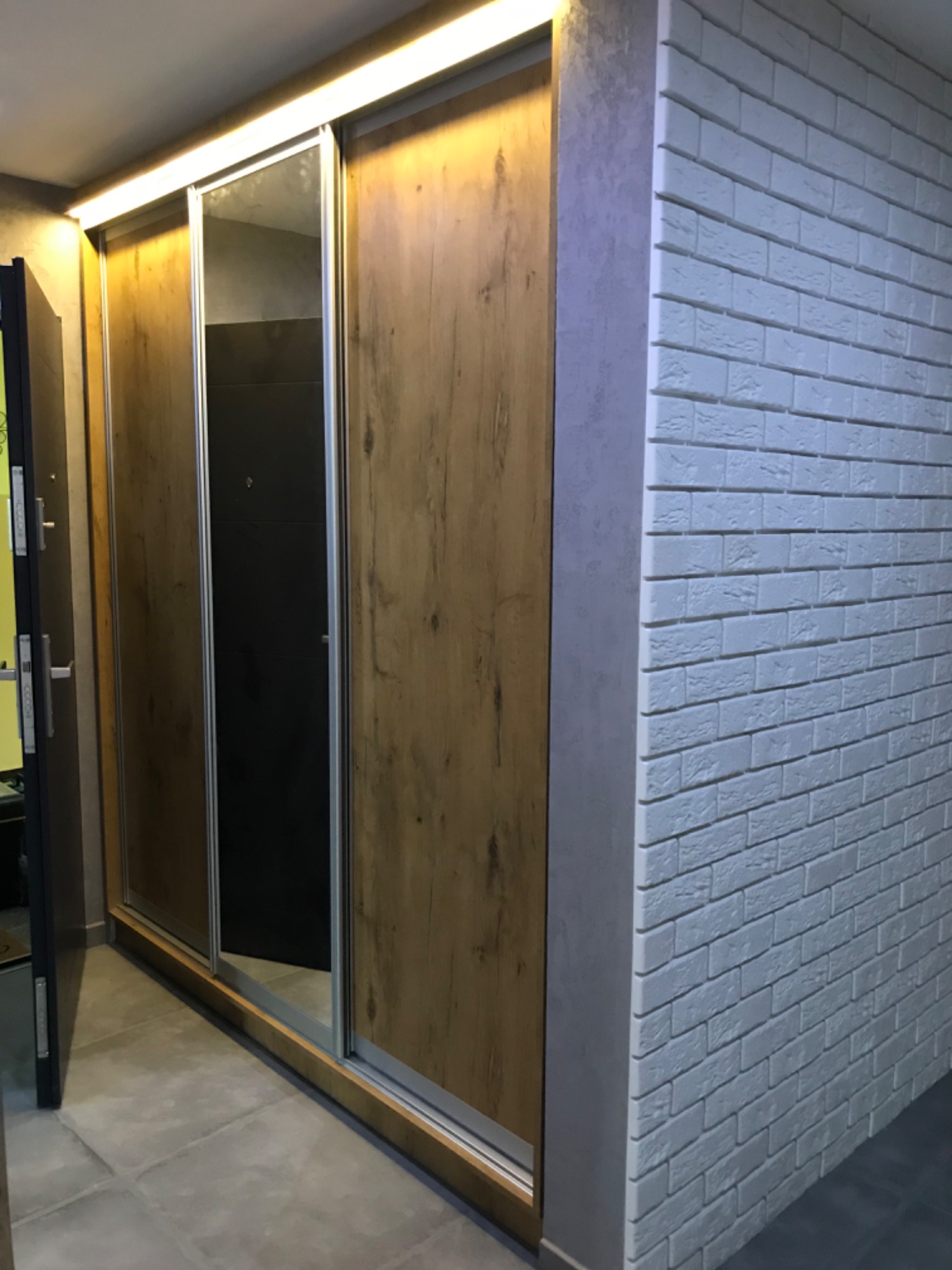 Szafa wnękowa z przesuwnymi drzwiami, wykończona drewnem i lustrem, z oświetleniem LED u góry, przylegająca do ściany z białej cegły.