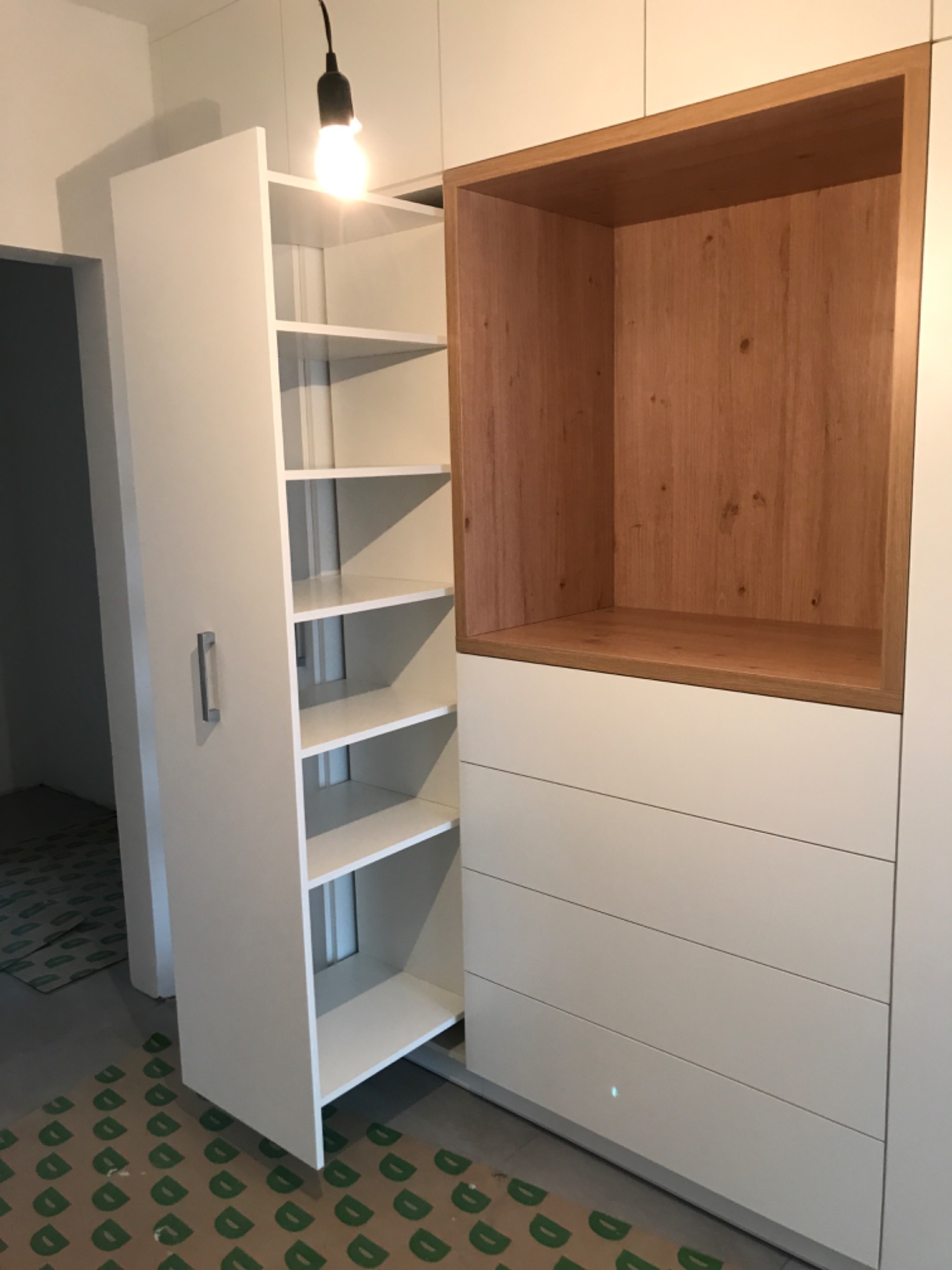 Biała szafa wnękowa z otwartą częścią z półkami i szufladami, wykończona drewnianym elementem, widoczna na podłodze folia ochronna.