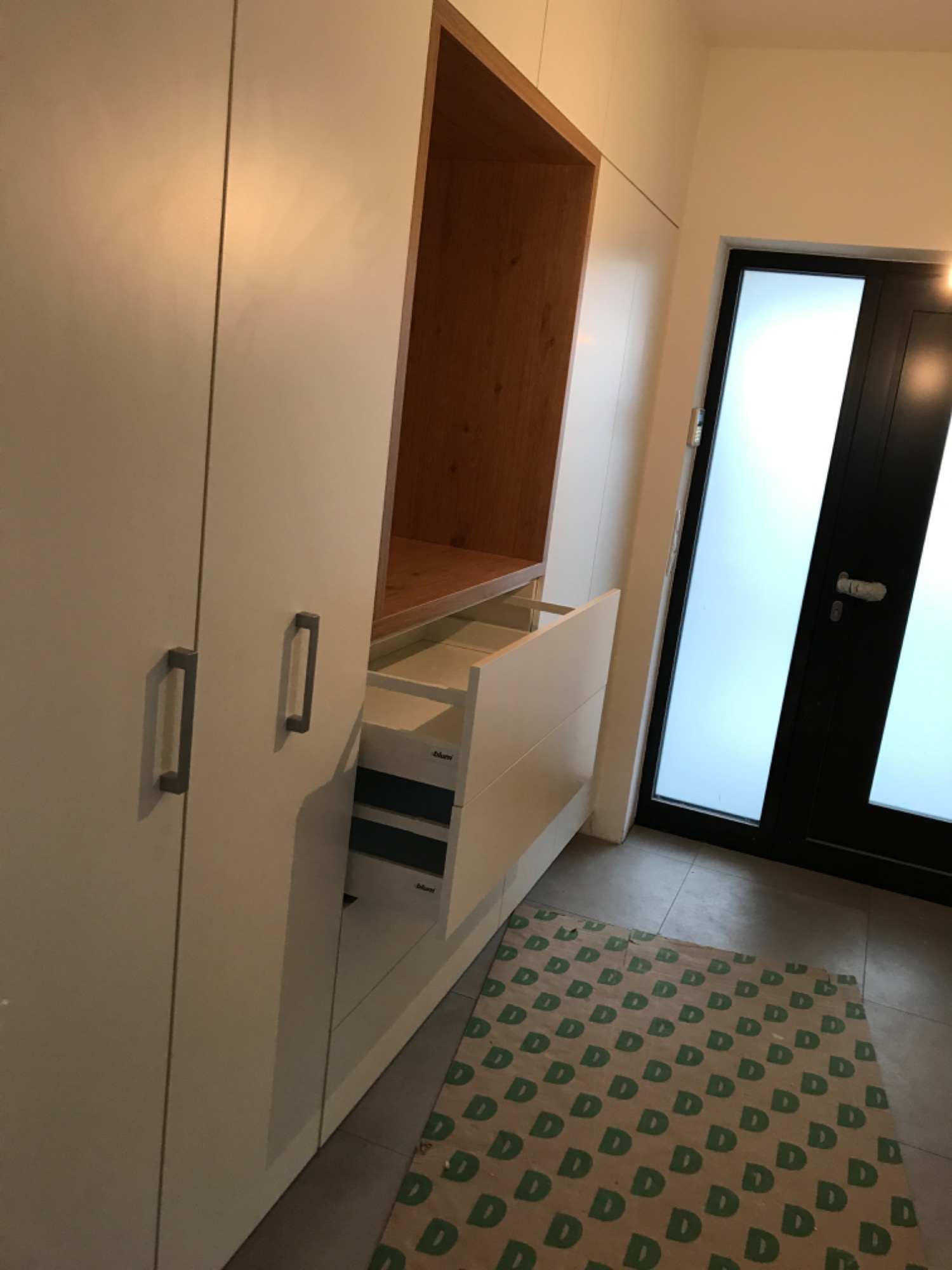 Biała szafa wnękowa z otwartymi szufladami i drewnianą wnęką, widoczna mata podłogowa z wzorem litery 'D'.