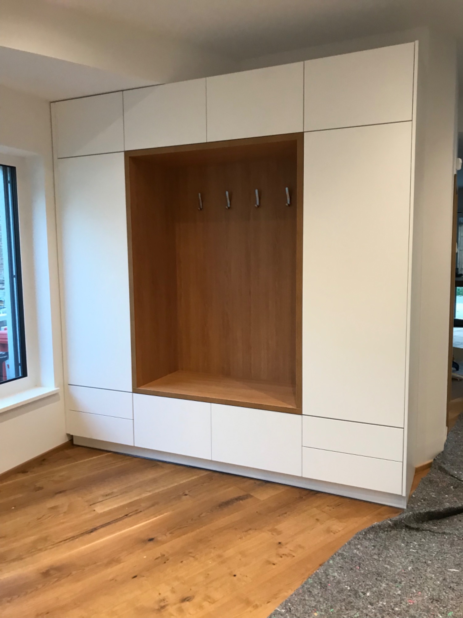 Biała zabudowa wnęki z drewnianym wykończeniem i haczykami na ubrania, z widoczną podłogą z jasnego drewna.