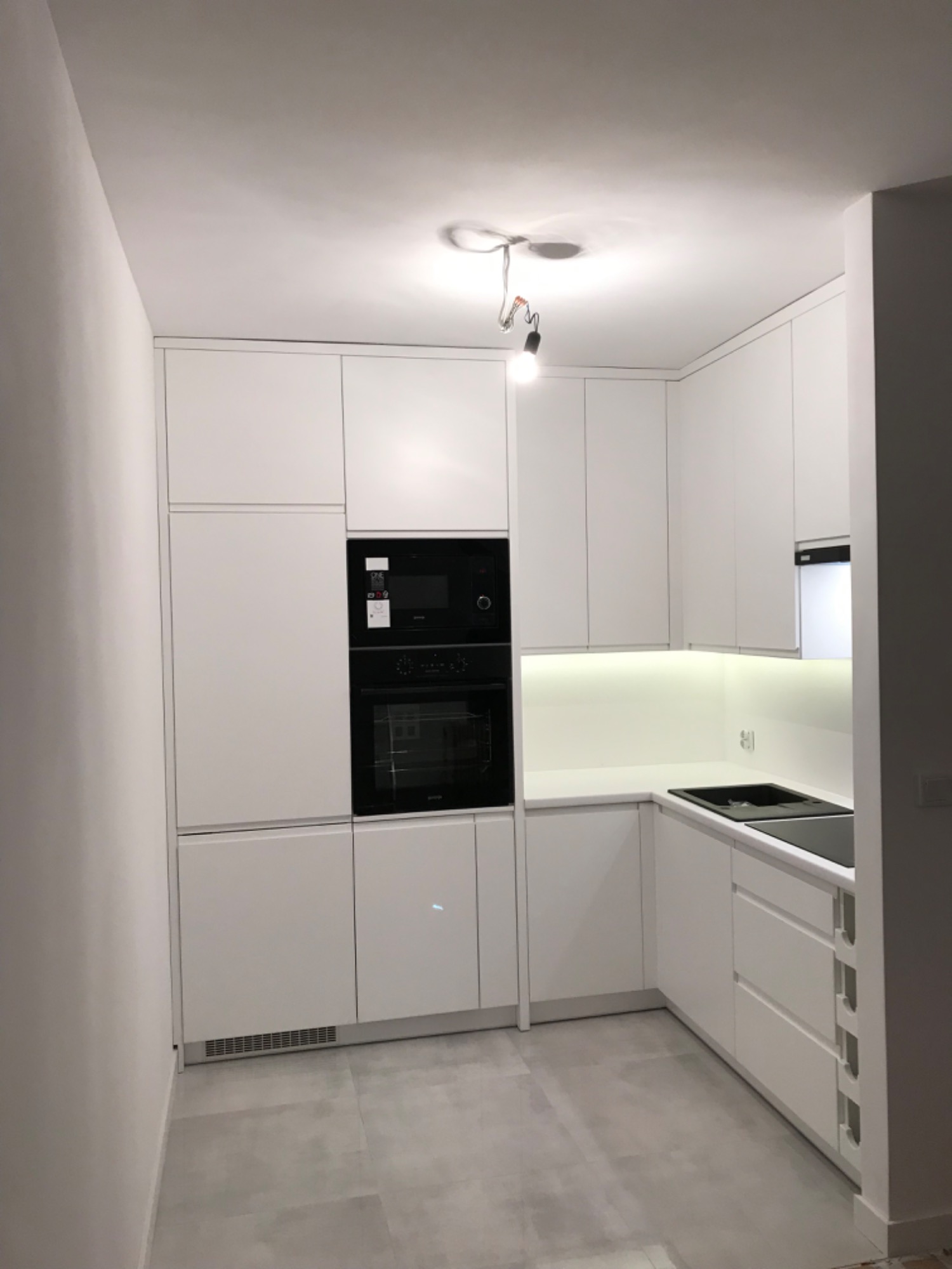 Nowoczesna, minimalistyczna kuchnia w bieli z zabudowanymi sprzętami AGD i podświetleniem LED pod szafkami wiszącymi, widoczne niedokończone podłączenie oświetlenia sufitowego.