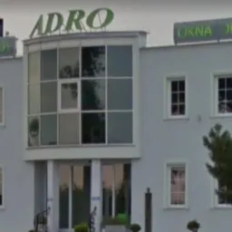 Budynek firmy ADRO z dużymi oknami wystawowymi i szyldami reklamowymi 'Drzwi Fasady' oraz 'Okna Drzwi' na dachu.