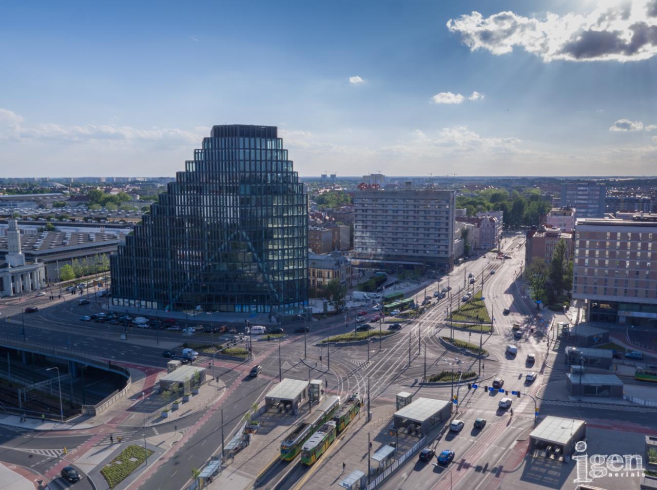 Panoramiczny widok z lotu ptaka na nowoczesny biurowiec o nietypowej, schodkowej architekturze, z tramwajami i ruchem ulicznym w dole, w słoneczny dzień.