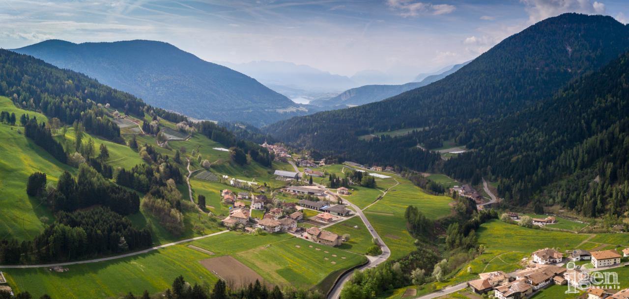 Panoramiczny widok z lotu ptaka na malowniczą wioskę w dolinie otoczonej zalesionymi wzgórzami, z polami uprawnymi i krętą drogą prowadzącą przez osadę, w tle majaczą odległe pasma górskie...