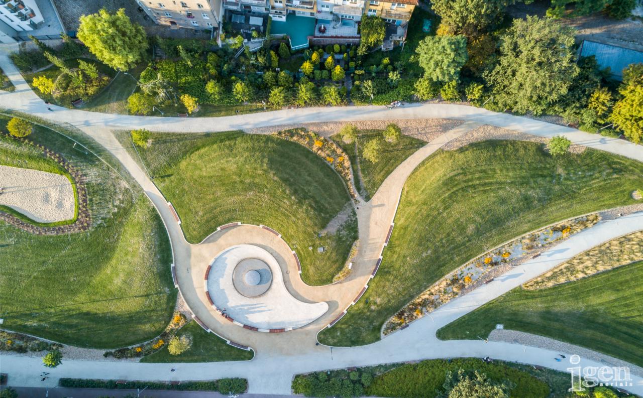 Widok z lotu ptaka na park miejski z alejkami, trawnikami, rabatami kwiatowymi i centralnym placem z ławkami.