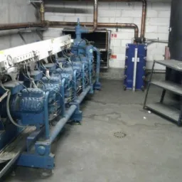 Szereg niebieskich sprężarek przemysłowych połączonych rurami miedzianymi w pomieszczeniu technicznym z betonowymi ścianami i podłogą.