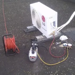 Biała jednostka zewnętrzna klimatyzatora LG na dachu, podłączona do pompy próżniowej i manometru, widoczne narzędzia i zwinięty kabel.