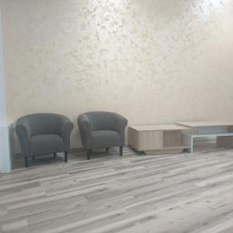 Świat Komfortu - Jasne panele podłogowe ułożone wzdłuż pomieszczenia, dwa szare fotele i stoliki kawowe na tle ściany z efektem dekoracyjnym.