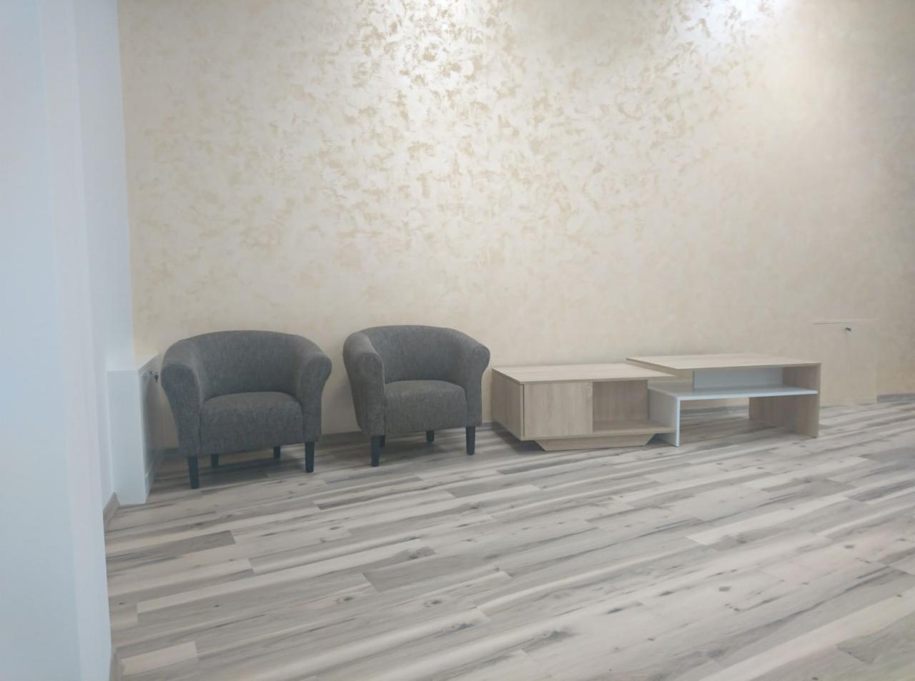 Jasne panele podłogowe ułożone wzdłuż pomieszczenia, dwa szare fotele i stoliki kawowe na tle ściany z efektem dekoracyjnym.