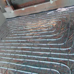 Rozłożona instalacja ogrzewania podłogowego na folii z siatką, rury przymocowane czarnymi uchwytami, widoczne logo SIGMA-LiS na rurach.